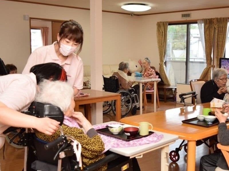 テーブルを囲んで食事をしている入居者たち。一目に高齢女性のそばにポロシャツを着た女性スタッフが2名付き添っている。
