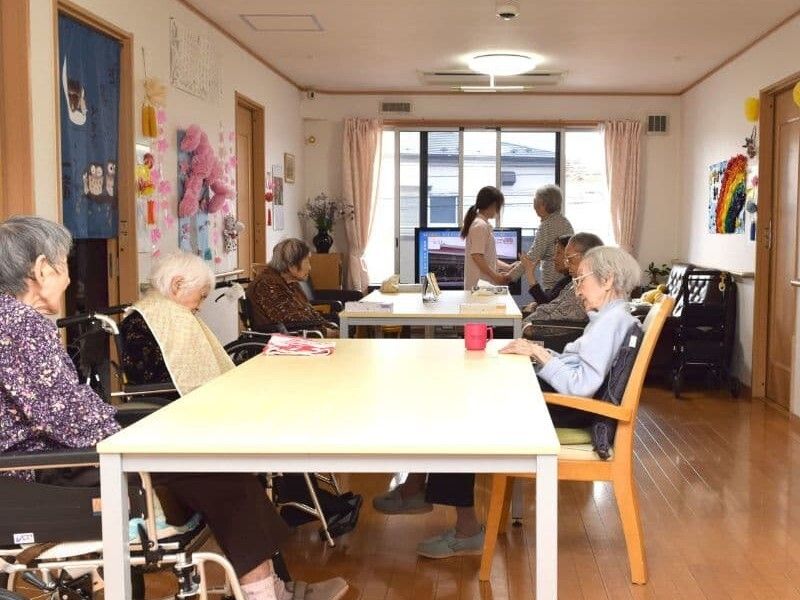 大きなダイニングテーブルセットが置かれ、車いすの利用者を含め入居者がテーブルを囲んでいる。奥には入居者の歩行を手伝うスタッフがいる。
