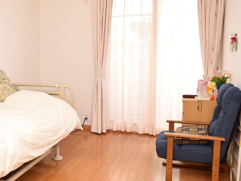 大きな窓が付いたフローリングの居室には寝具付きのベッドやいす、タンスなどがある。