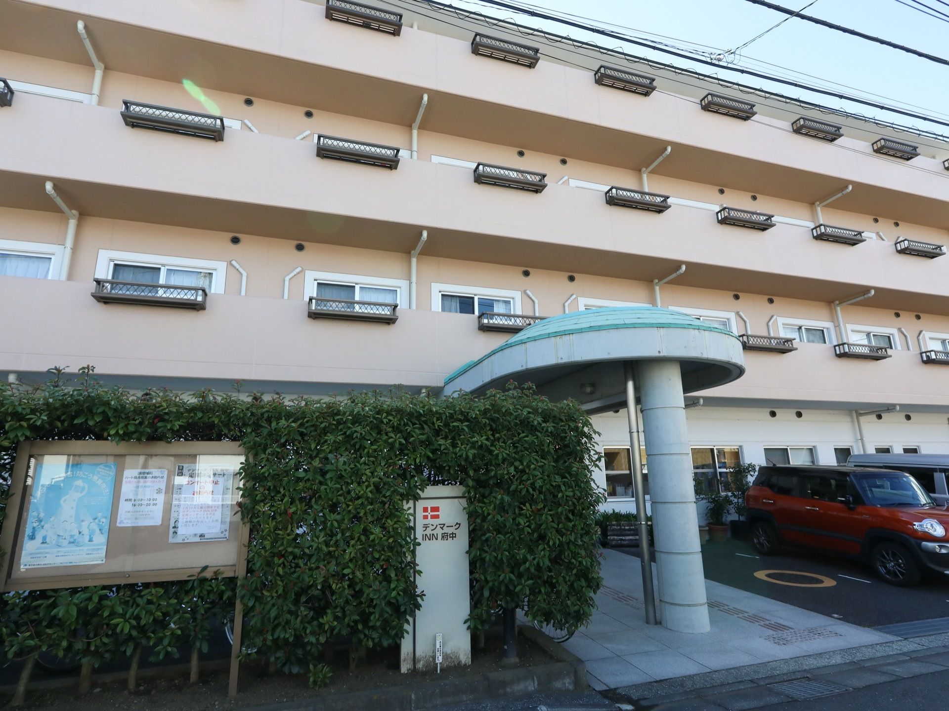 生垣に施設の看板が立っており、その脇に掲示板が設置されている。玄関の屋根が施設入口まで張り出している。