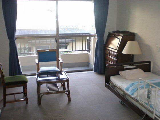 大きな窓が付き明るい雰囲気の居室には、寝具付きのベッドやいすなどがたくさんあり、フロアカーペットが敷かれている。