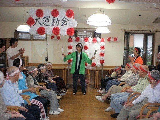 紅白のハチマキを付けた入居者がスタッフの説明を聞いている様子。「大運動会」と書かれた看板が掲げられ、スタッフはハッピを着ている。