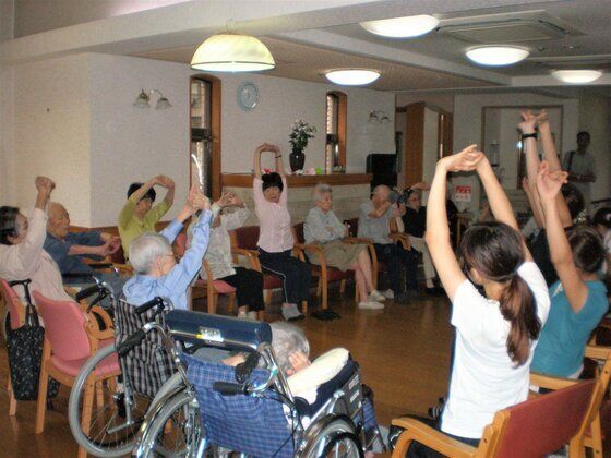 椅子に座って両手を上に上げている入居者とスタッフ。車いすに座っている入居者も参加している。