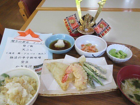 たけのこご飯や天ぷらの盛り合わせ、そら豆などの野菜の小鉢、かきたま汁などが並び、兜の置物とお品書きも置かれている。