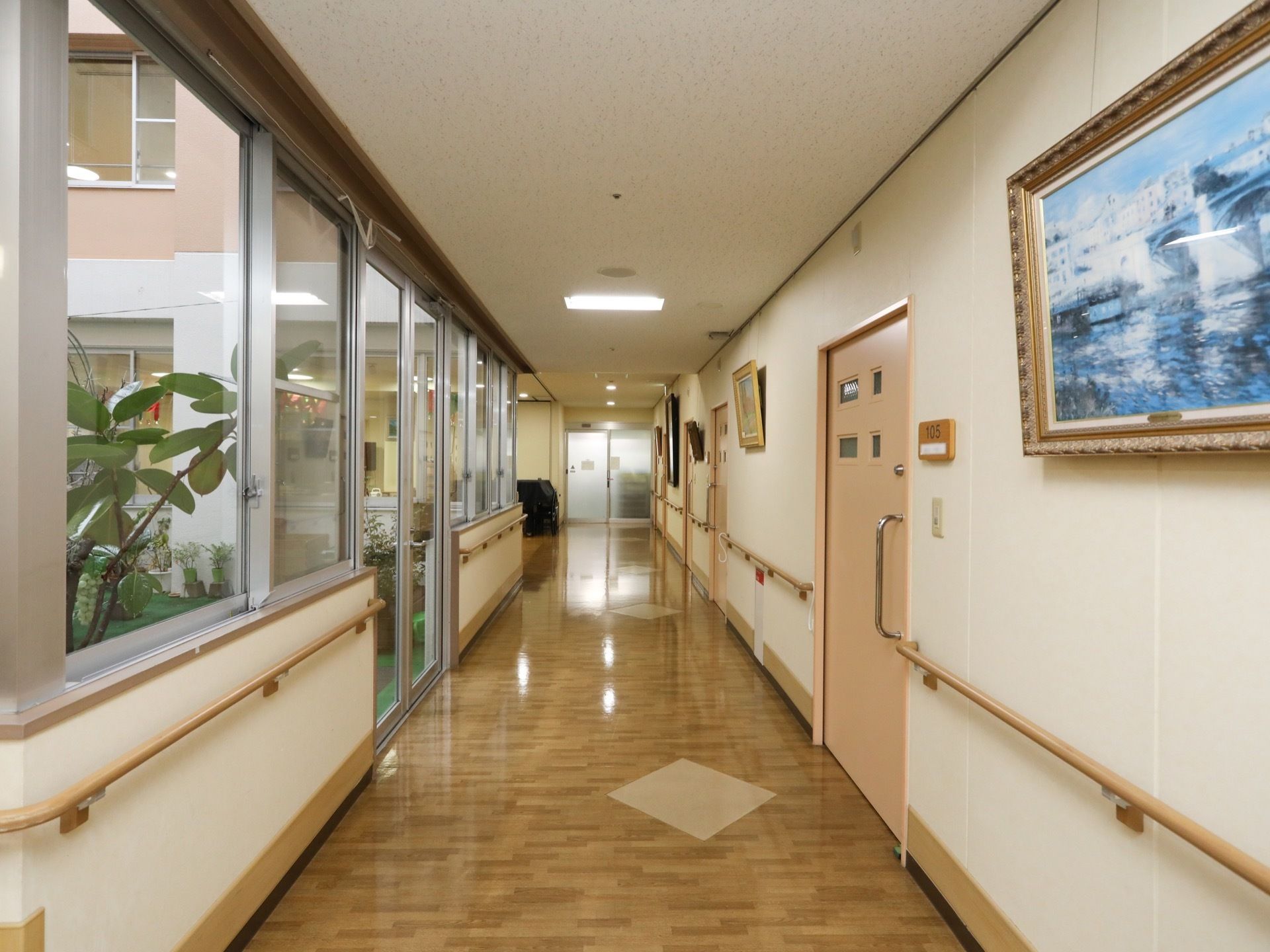 木目調の廊下が伸びている。片側には居室の入り口が並び、反対側は中庭に面した窓になっている。壁には手すりが付いている。