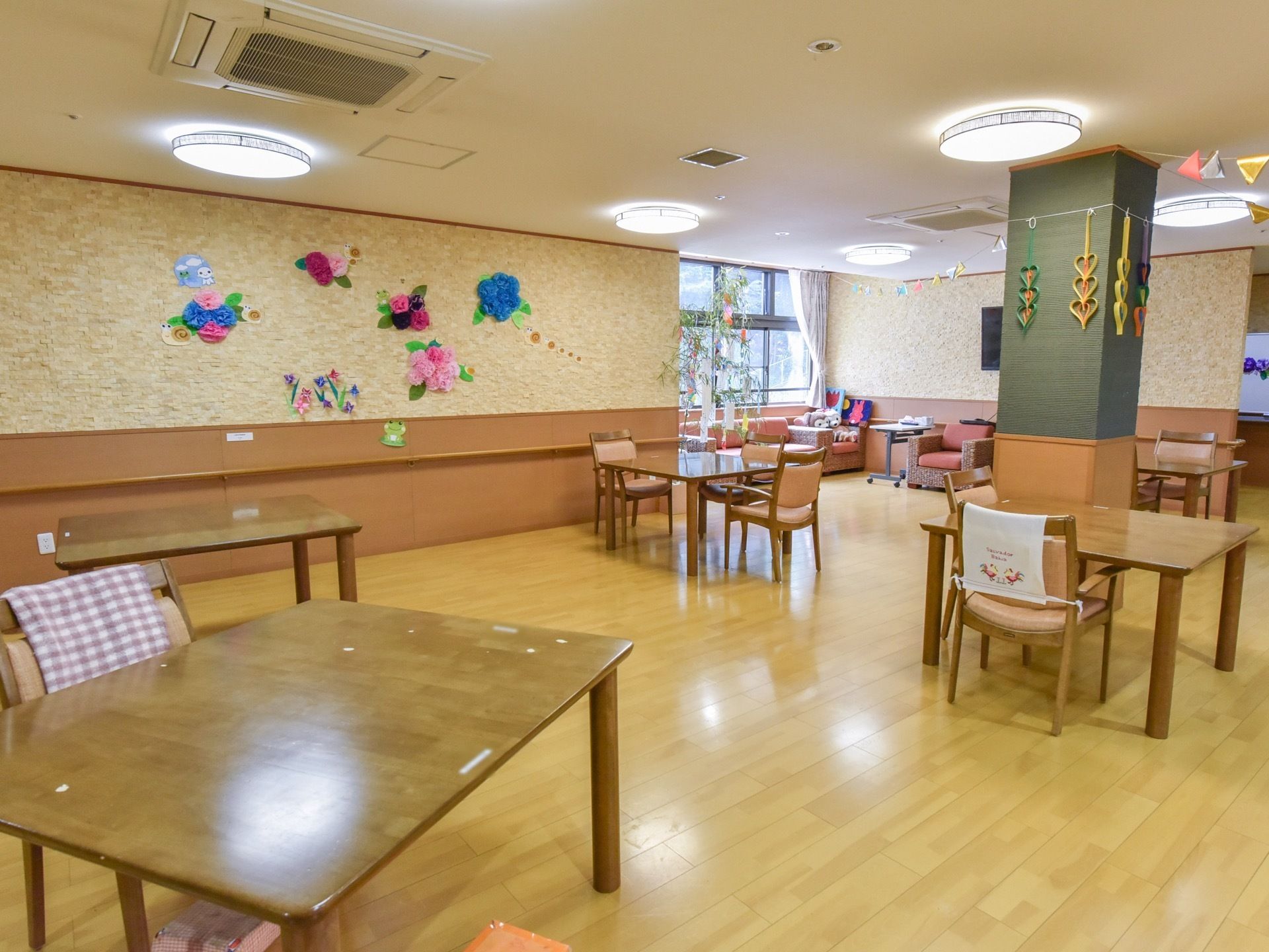 フローリングの床の広い部屋になっており、椅子とテーブルが置かれている。柱や壁には折り紙などの作品が飾られている。