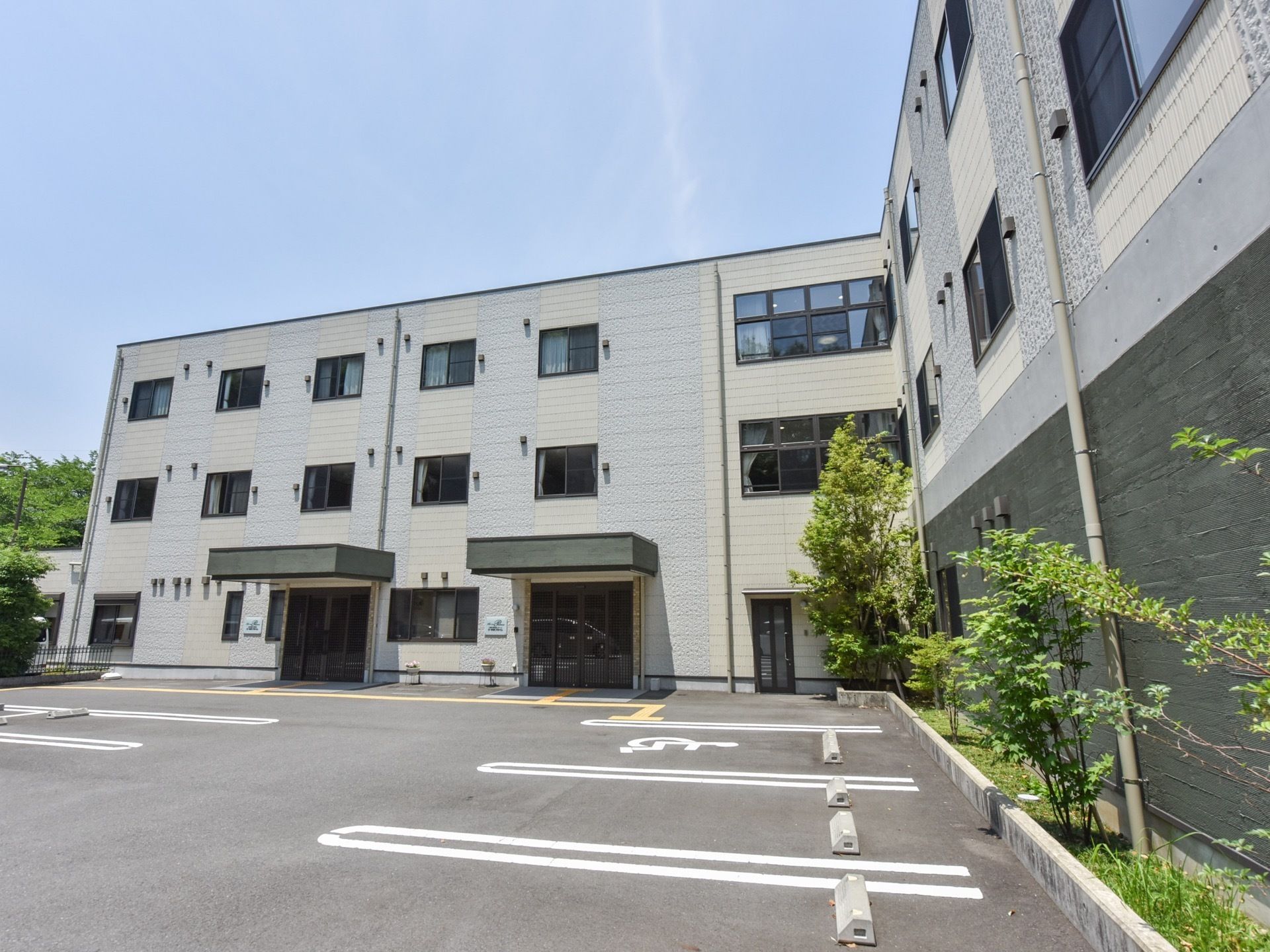 施設はL字型の建物になっており、各居室の窓がみえる。施設を囲むようにさまざまな大きさの木が植えられている。