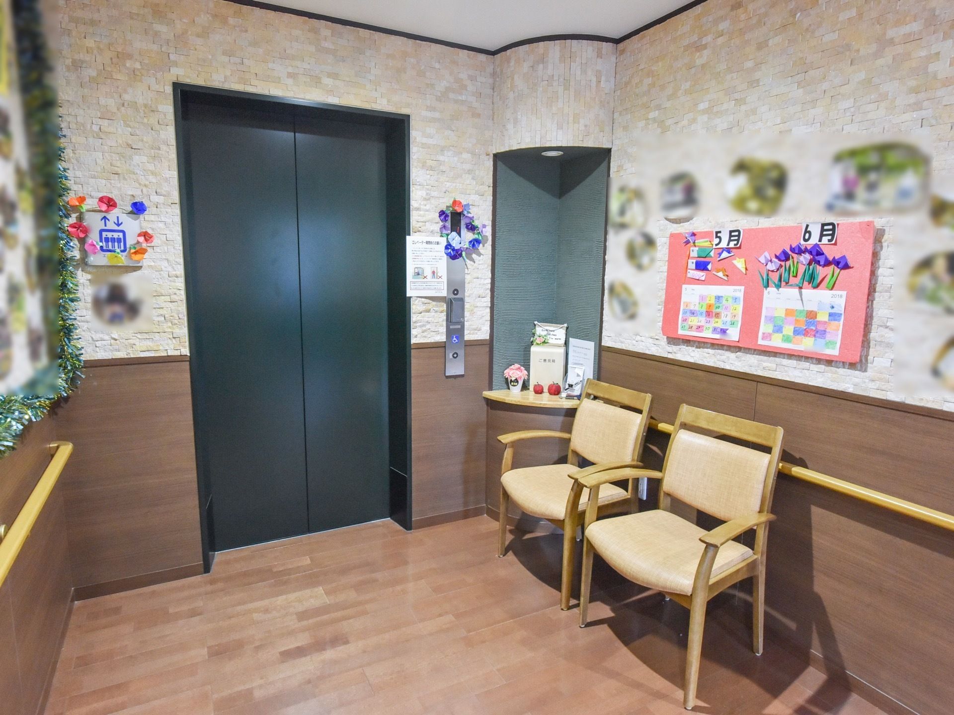 エレベーター前に椅子が2脚用意されている。壁には手作りのカレンダーや写真などの掲示物が飾られている。