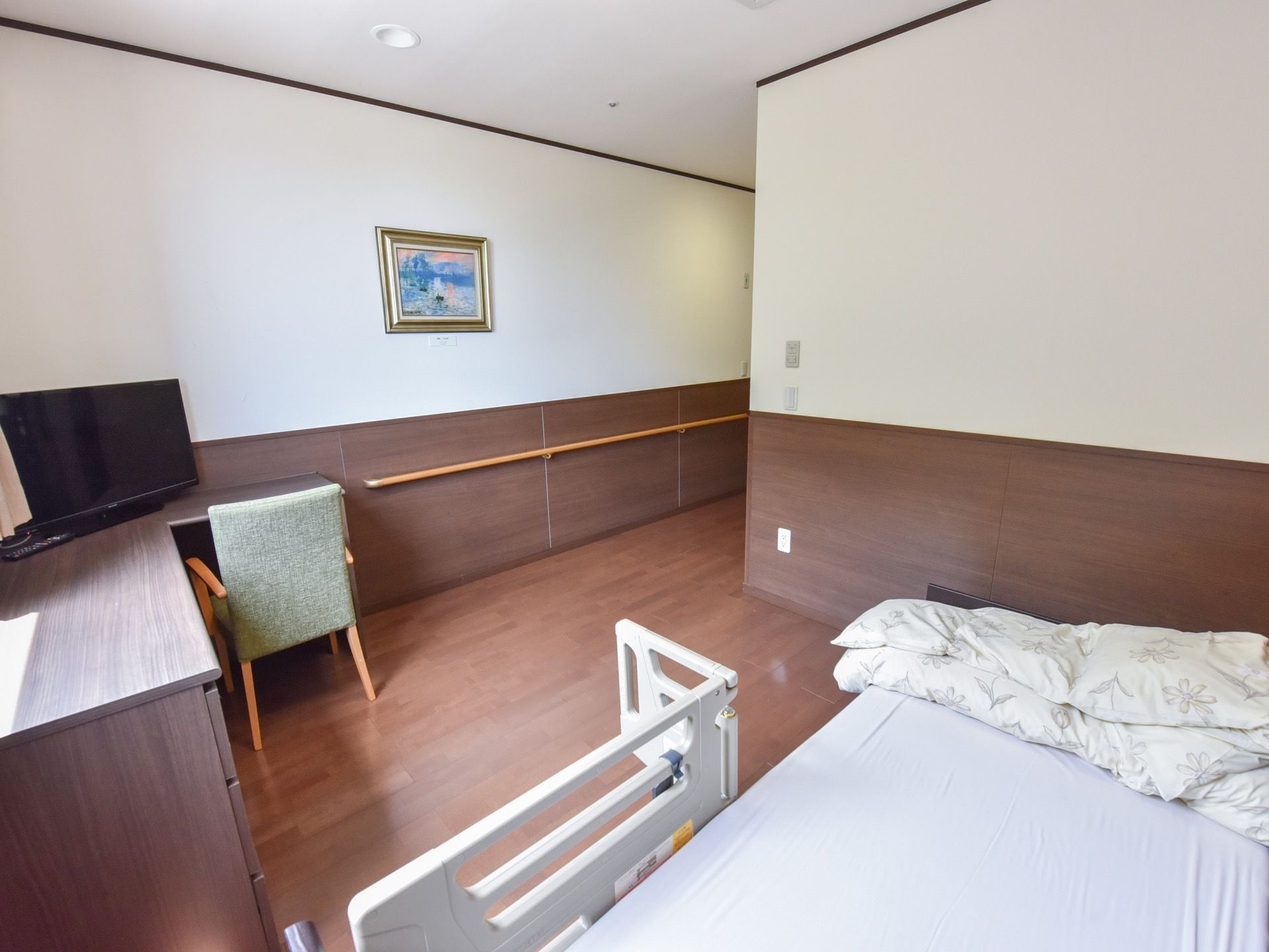 壁と色を揃えたデスクの上にテレビが置かれ、その前に椅子が1脚用意されている。壁には絵画が飾られている。