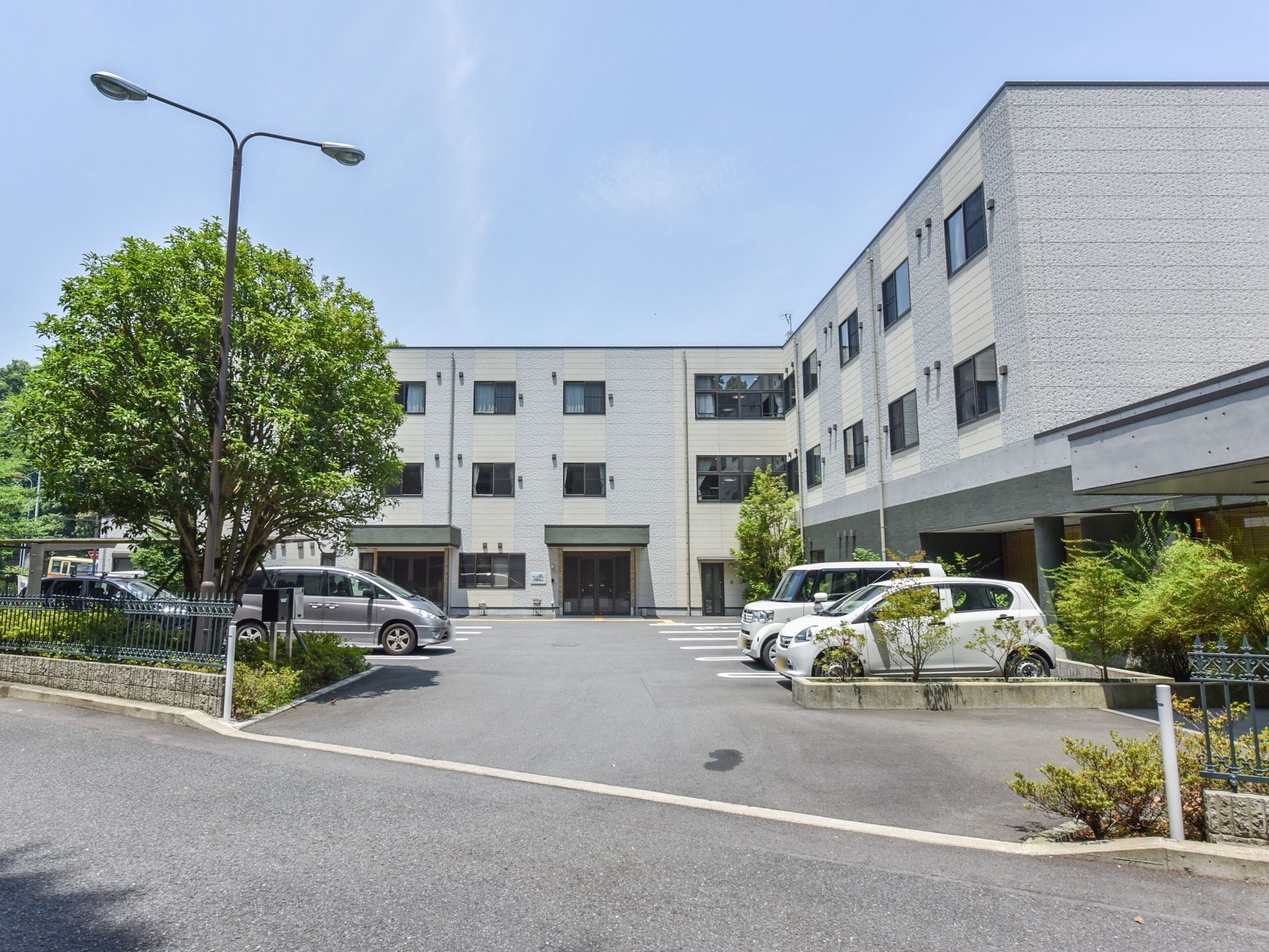 地上３階建の施設は、ライトグレーを基調とした上品なデザインである。前方には広い駐輪場があり、左端には大きな木がある。