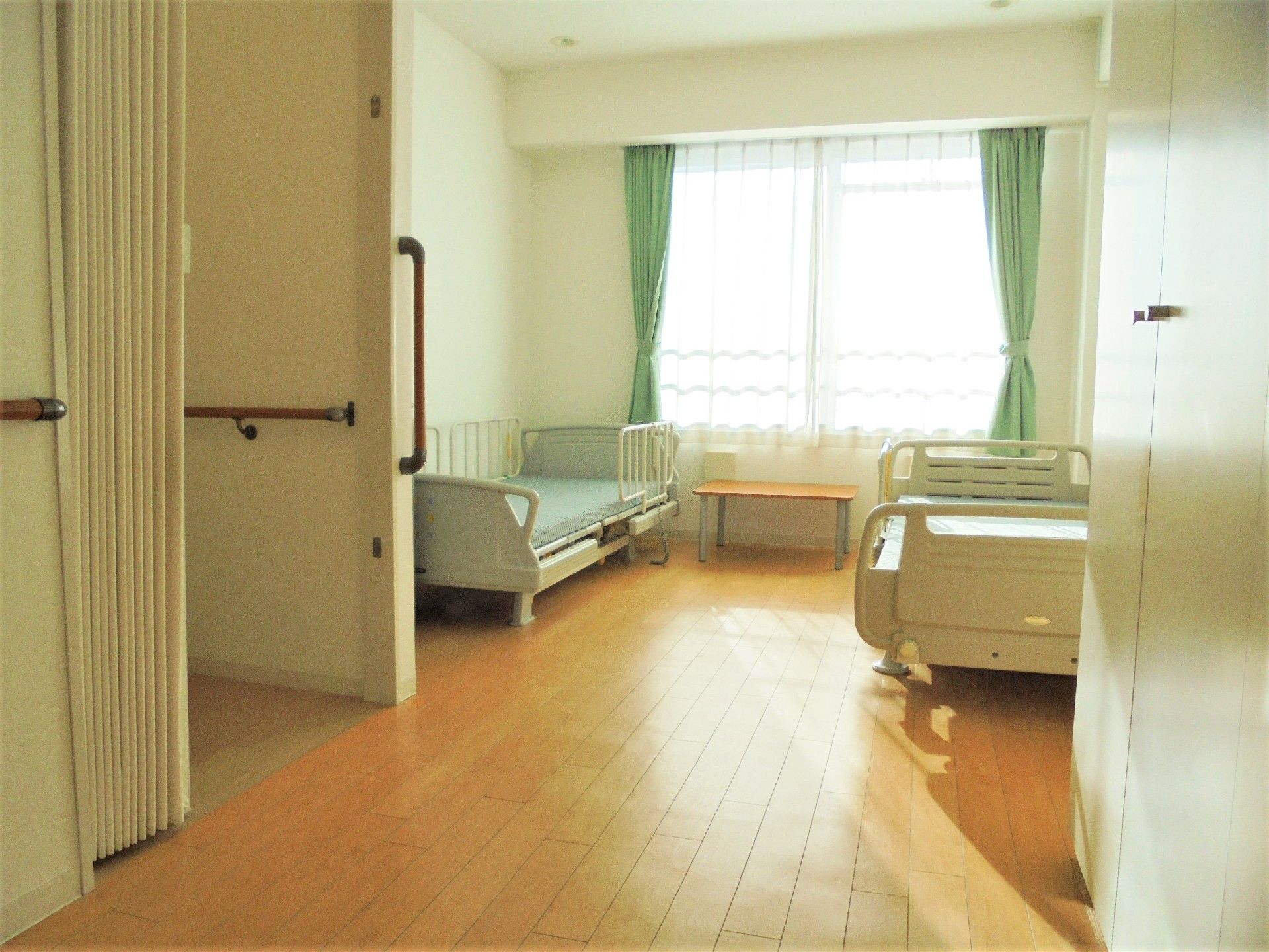 ベッドが二つ置いてある二人部屋の様子。明るい日差しが差し込む窓と手前にトイレの様子
