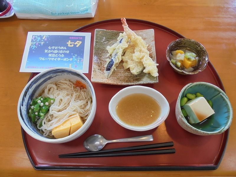 半月型のお盆に乗っている行事食。うどんやてんぷら、そのほかの小さなおかず類が並んでいる様子