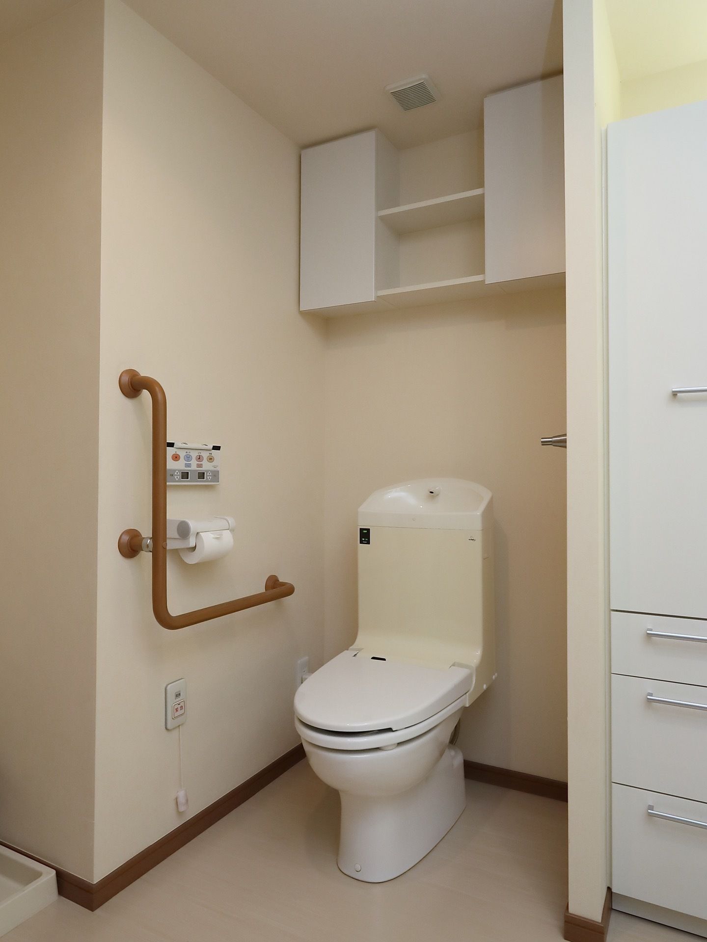 居室にトイレが完備されている。壁には温水洗浄機能の操作盤、緊急ボタン、手すりが取りつけられている。便座後ろの上には収納棚が設置されている。