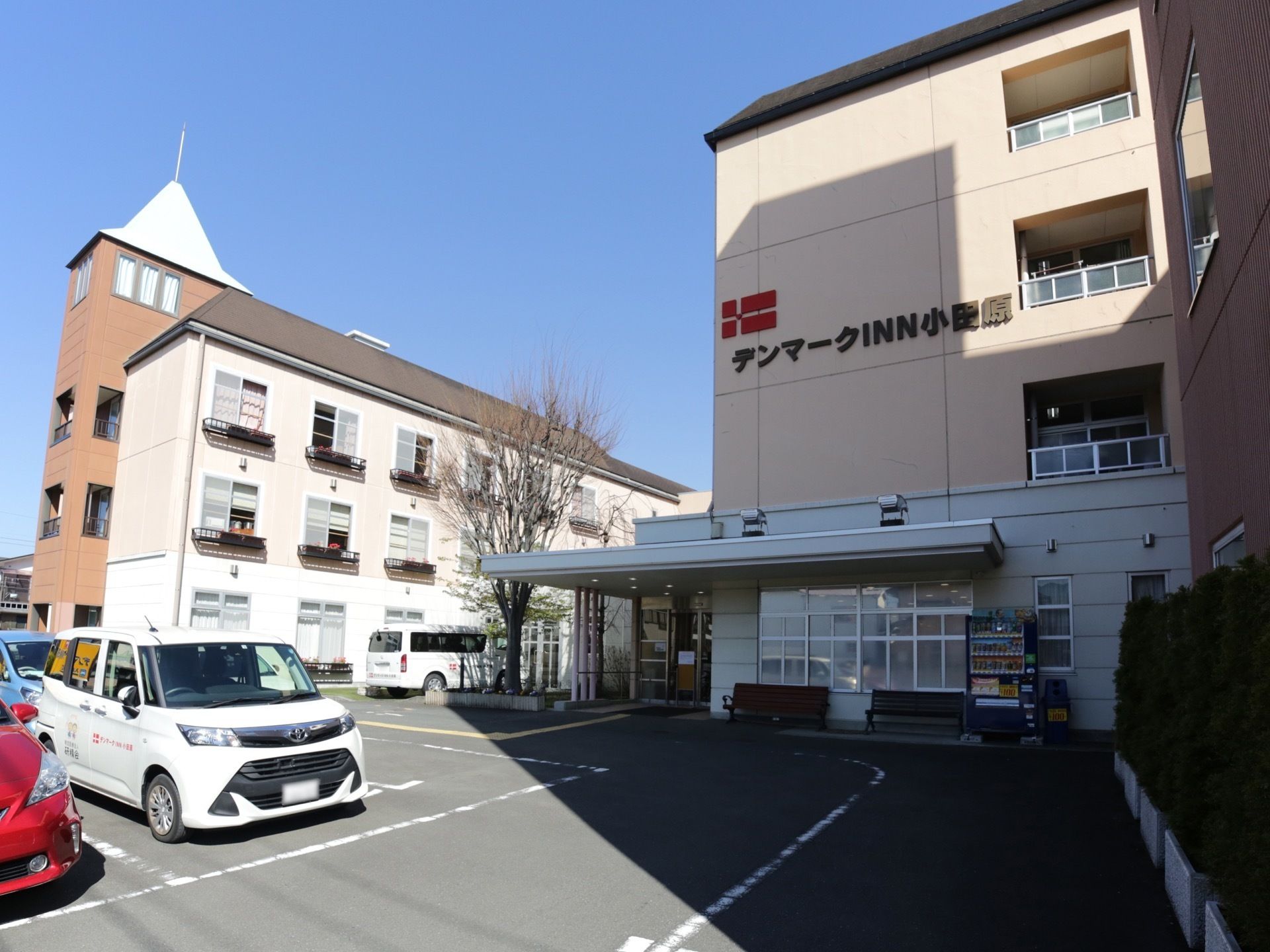 アスファルトの駐車場があり、奥に４階建ての大きな施設が建っている。玄関前にはベンチがあり、自動販売機も設置されている。