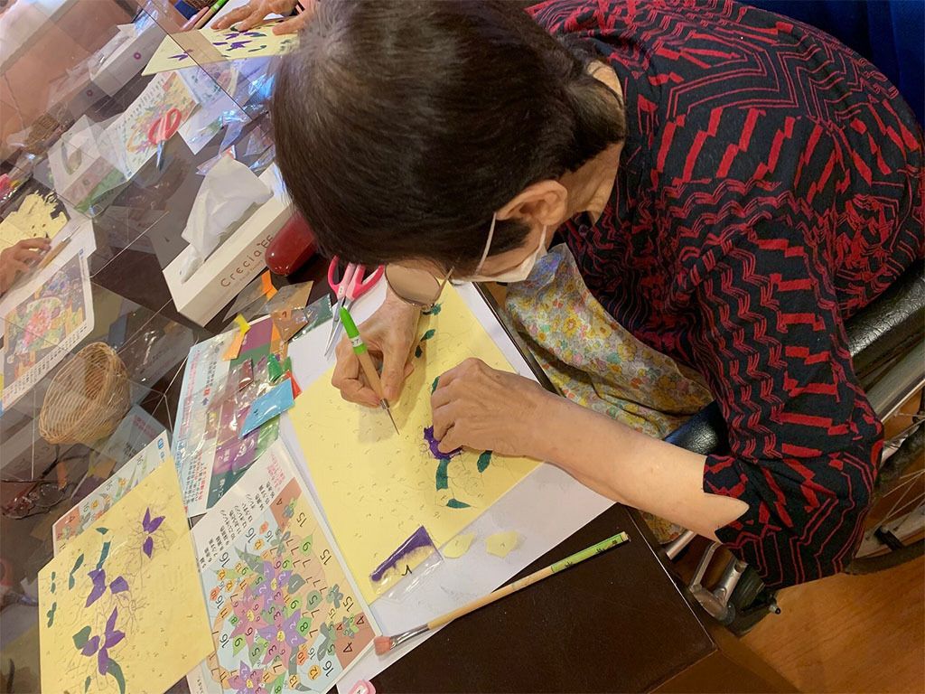 下書きがされた紙を切り取っている高齢女性。真剣な表情で創作活動に取り組んでいる。