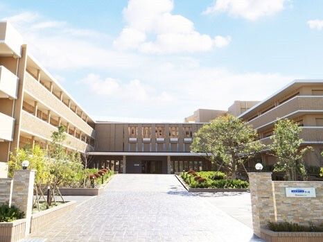 ベージュ系の３階建ての建物。玄関には屋根が付き、建物周辺には緑がある。立派な門にはホーム名が枯れたプレートが貼られている。