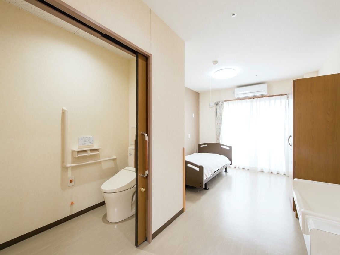 エル字型の手すりが付いた広いトイレがある居室には寝具付きのベッドが置かれ、エアコンも付いている。