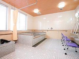 白い壁に薄いブルーのタイルが貼られたフロア、紫のシャワーチェアがあり、大小の浴槽が設置されている。