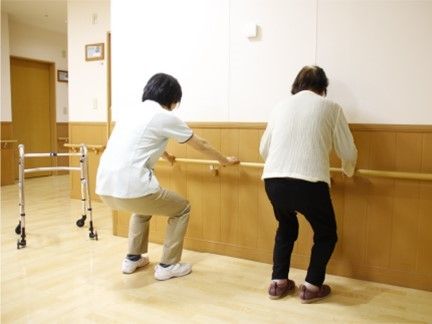 入居者とスタッフが廊下の手すりにつかまってスクワットをするときのように膝を曲げている様子。