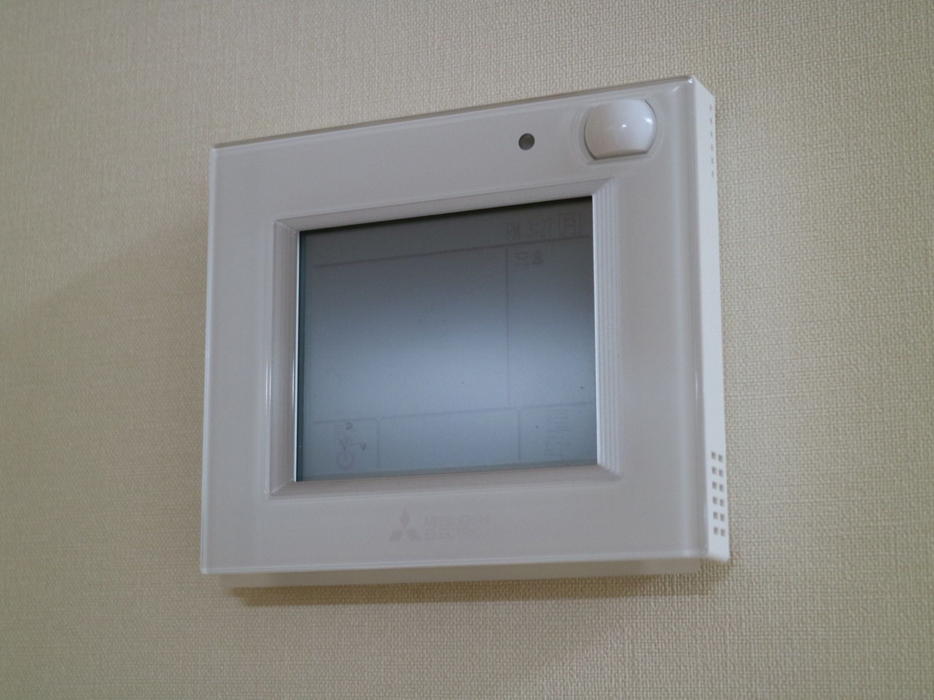 居室の壁面に、白色で四角い形をした、エアコンの操作パネルが設置されている。パネル中央には液晶画面が付いている。