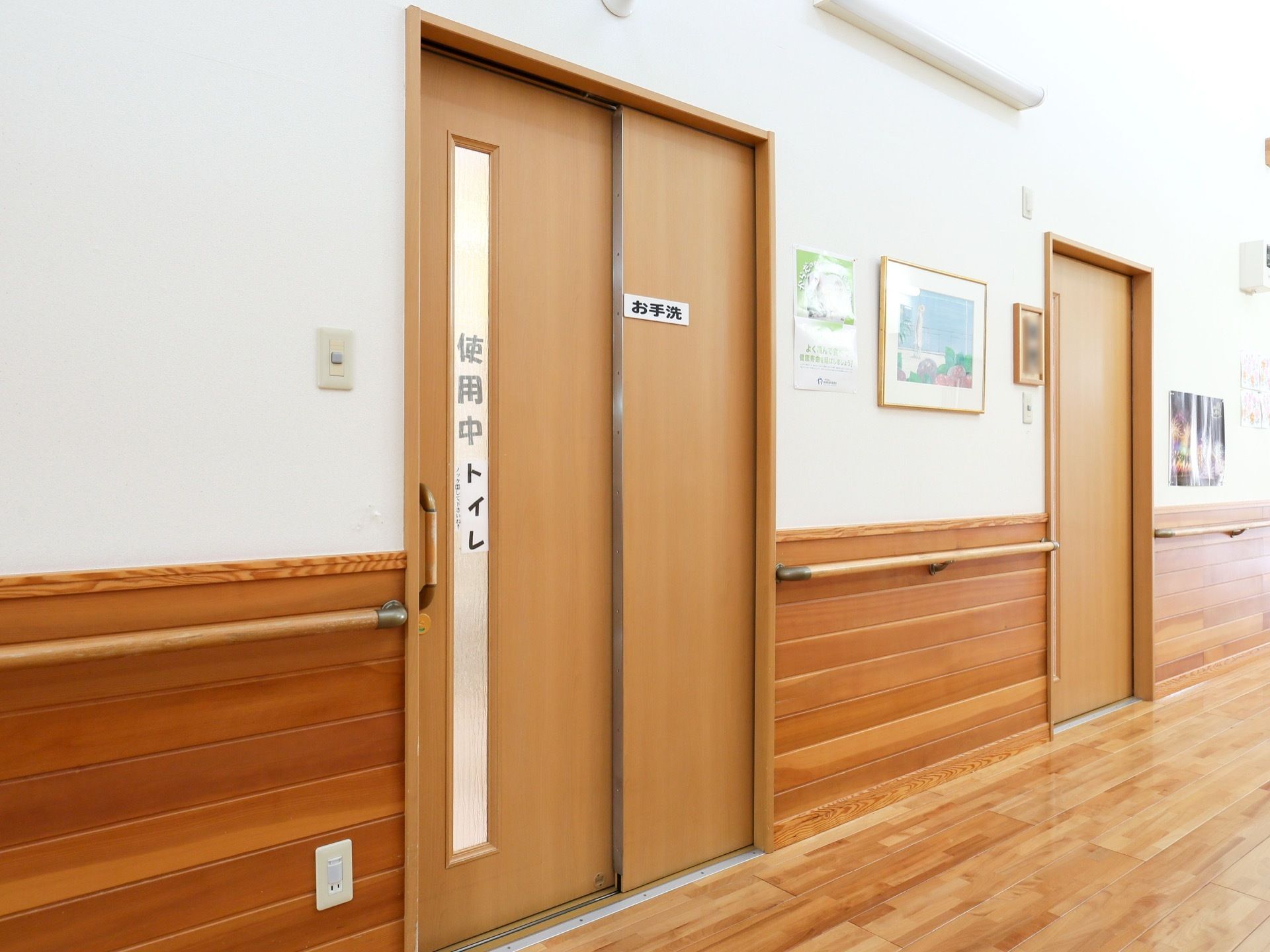 共用トイレの入り口。温かみの感じられる、木目調のスライドドアで、すりガラスには、使用中の文字が表示されている。