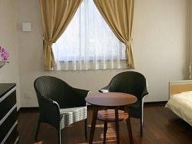 レースカーテンとカーテンが付いた窓がある部屋にはテーブルセットが置かれ、タンスやベッドもある。