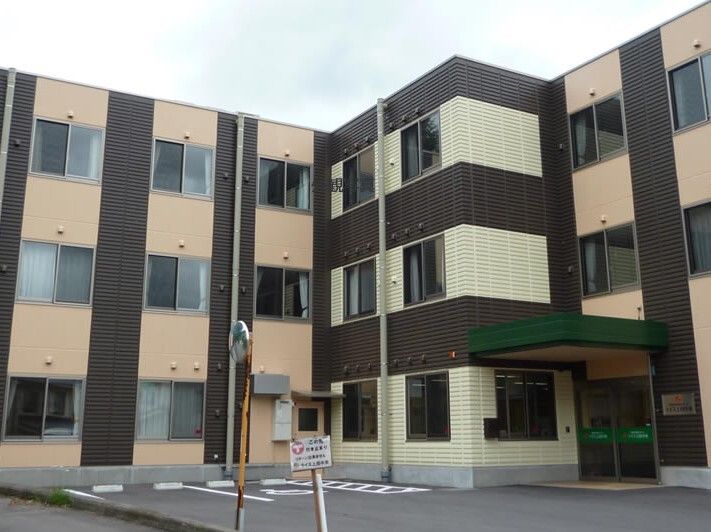 ベージュと黒の３階建ての建物はエル字型で玄関には屋根が付き、敷地内には駐車場があり、玄関には屋根がある。