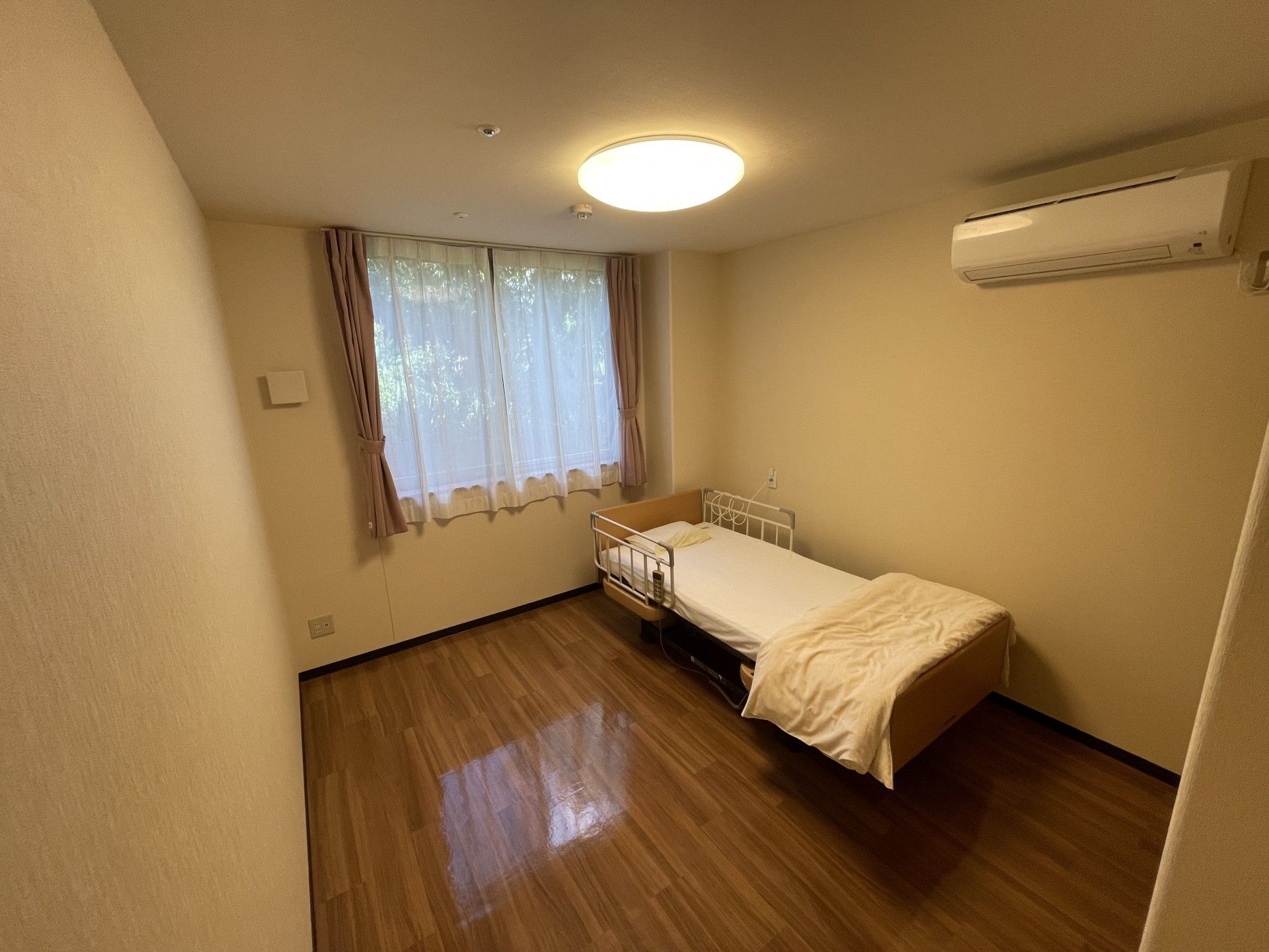 ベッドやテーブルセット、タンスなどが置かれたフローリングの部屋には、レースカーテンとカーテンが付いた窓もある。