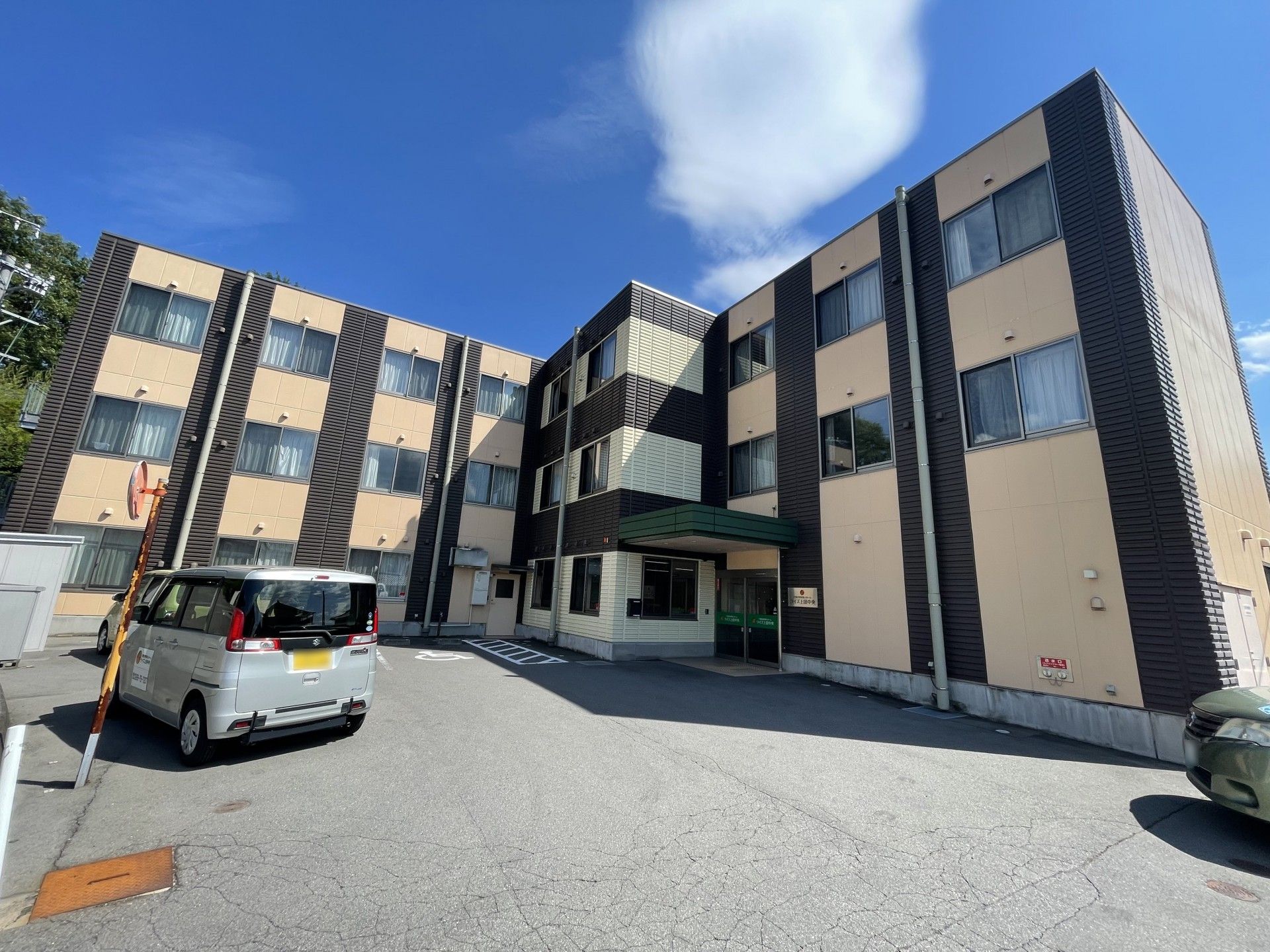 エル字型の３階建ての建物はベージュと黒で落ち着いた雰囲気。敷地内には駐車場があり、白線が引かれている。