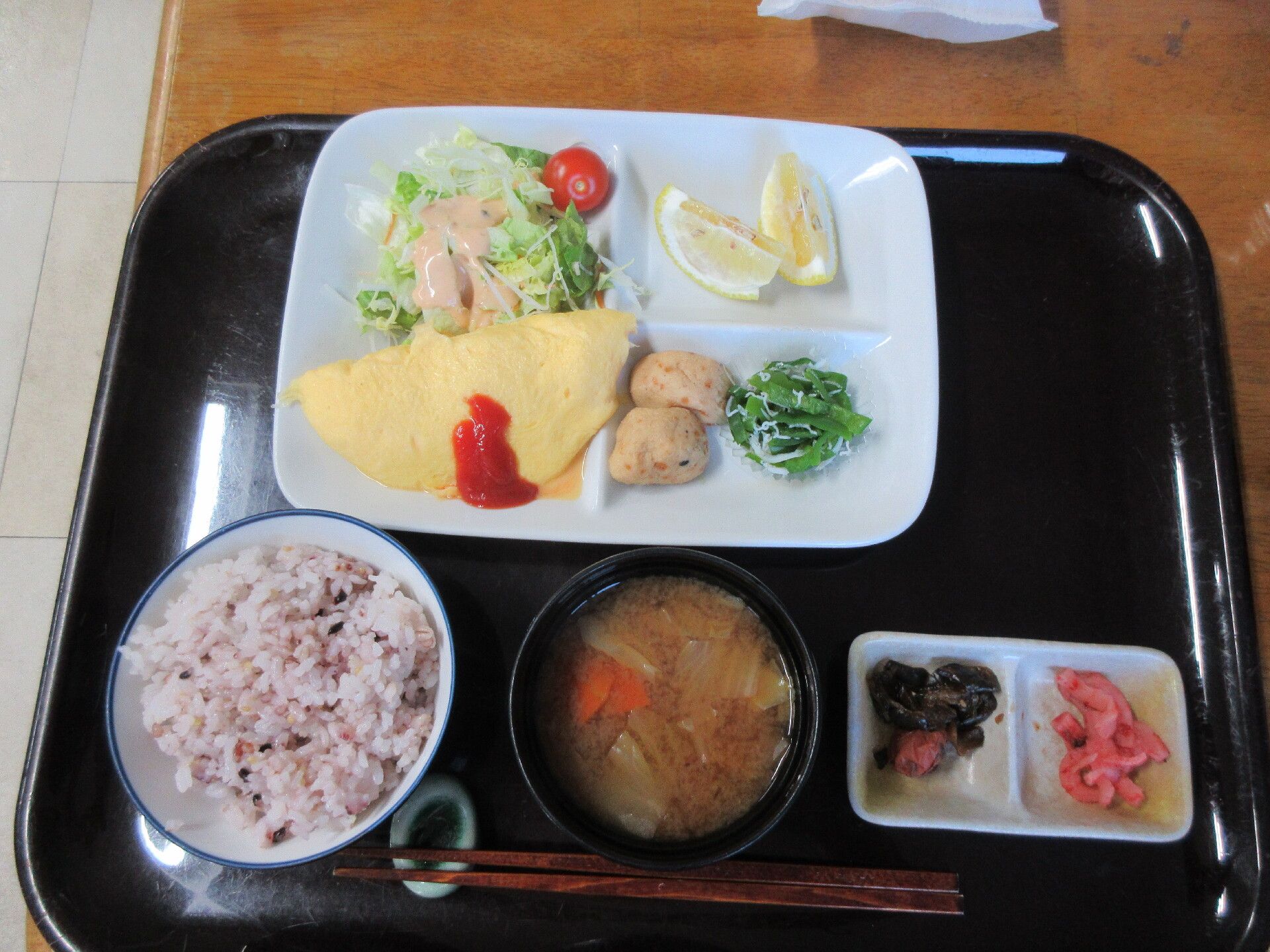 トレーの上には白いご飯とみそ汁、複数の煮物など野菜のおかずに焼き魚が並んでいる。