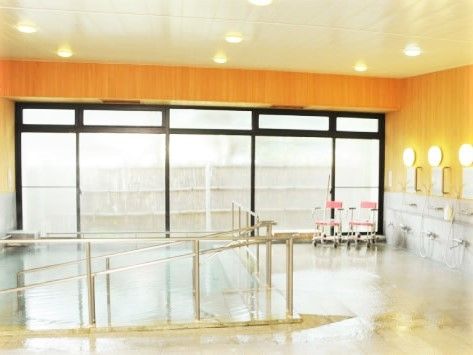 手すり付きのスロープが付いた大きな浴槽。右側にはシャワーが並び、浴室用の車いすもある。