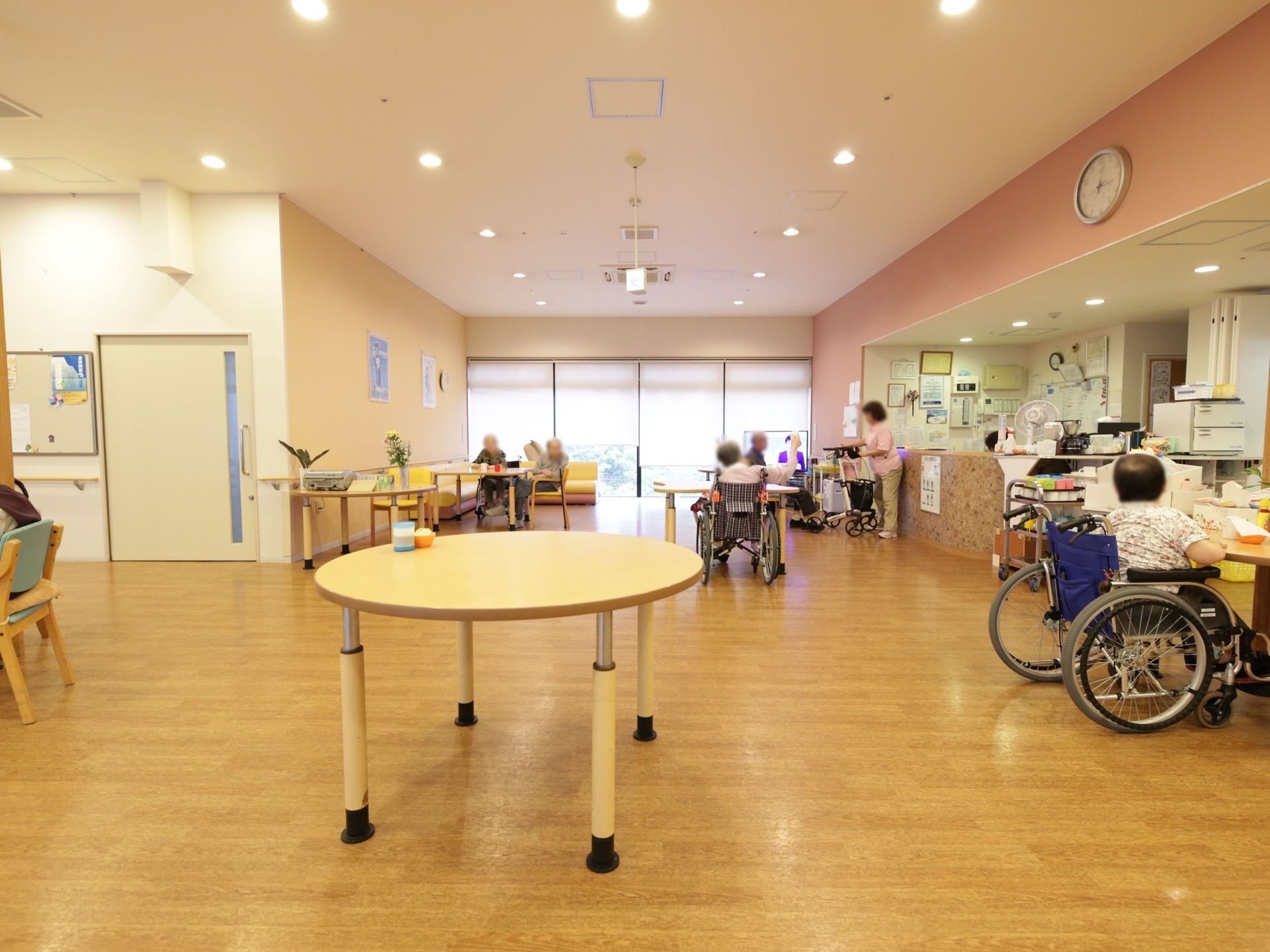 事務室の前が休憩スペースになっている。車椅子をご利用の入居者様が多く集まっている。女性スタッフが１名立っている。
