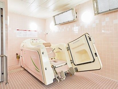 座ったまま入浴可能な機械浴を設置した浴室。白とピンクの車いす風の設備が付いた大きなマシンがある。