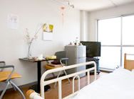 介護用ベッドの上にいる目線から部屋の中を写した様子。小さなテーブルと椅子、冷蔵庫、テレビなどがある