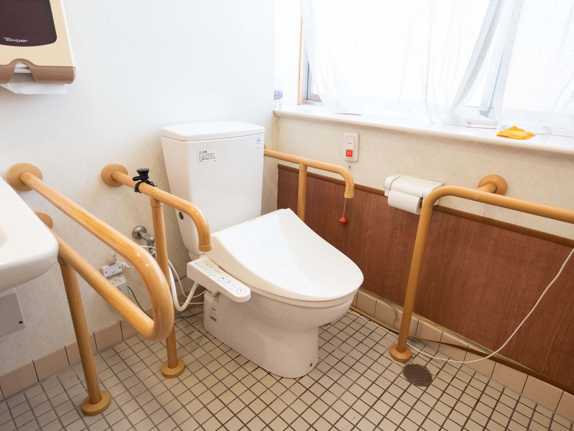トイレの窓にはレースカーテンが取り付けられている。手すりや緊急呼出ボタン、トイレットペーパーホルダーが設置されている。