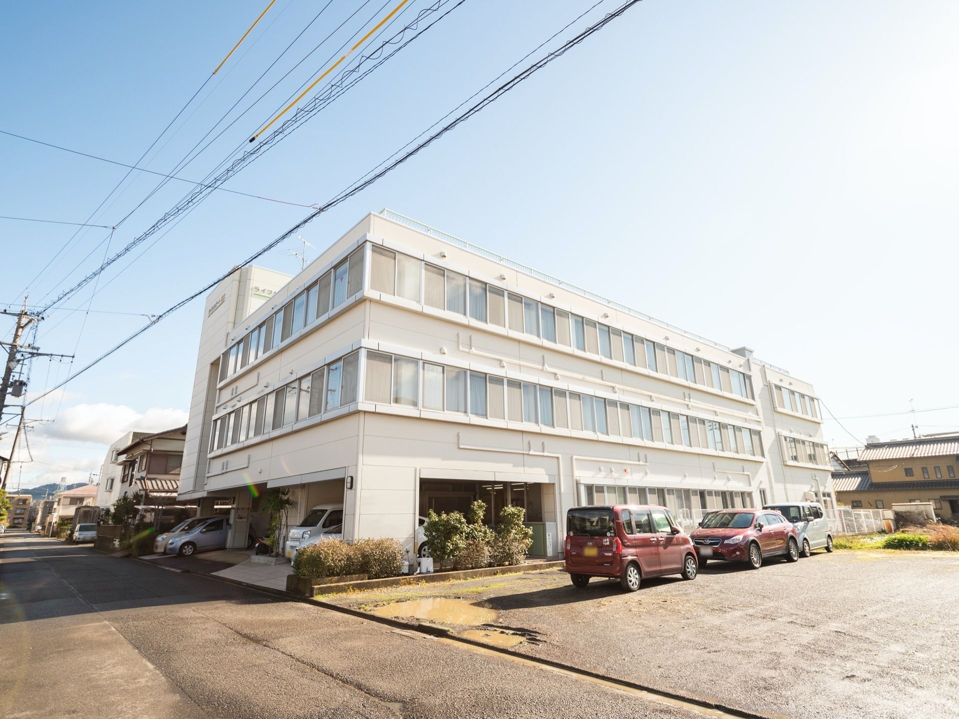 ３階建ての建物で、多くの窓が取り付けられている。外壁はホワイトである。いくつもの車が停められている。