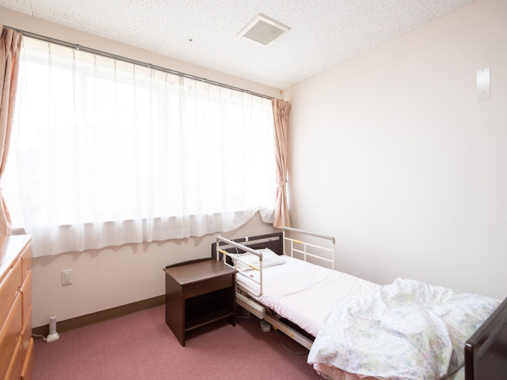 居室の窓にピンク系の遮光カーテンとレースカーテンが付けられている。ベッドとサイドテーブルが置かれている。