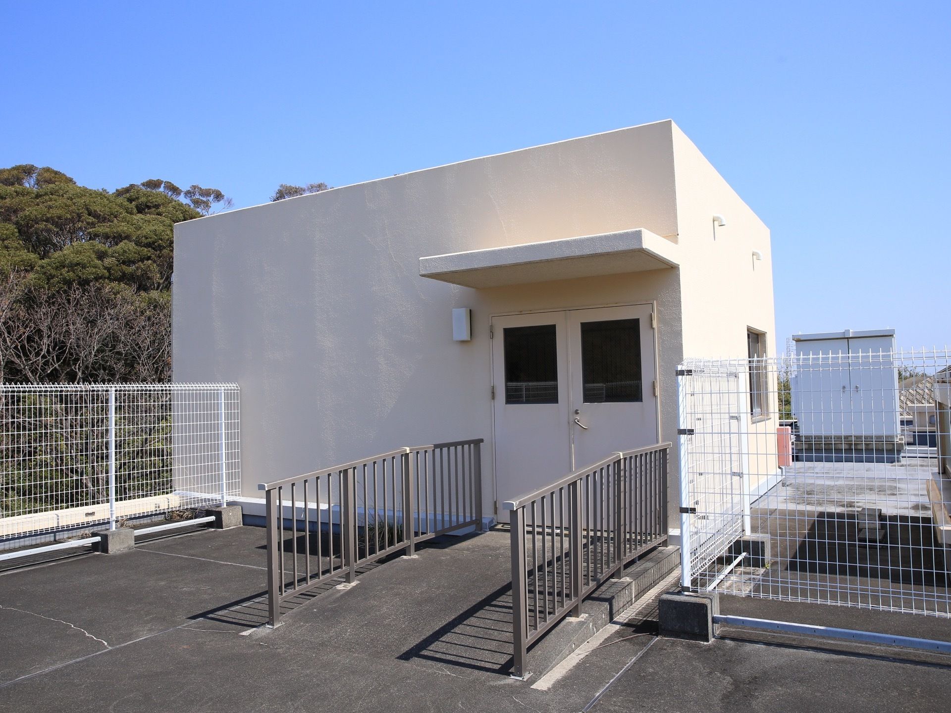 屋上に出る扉がある。扉の前は両側に柵がついたゆるやかなスロープになっている。周囲には白いフェンスが設置されている。