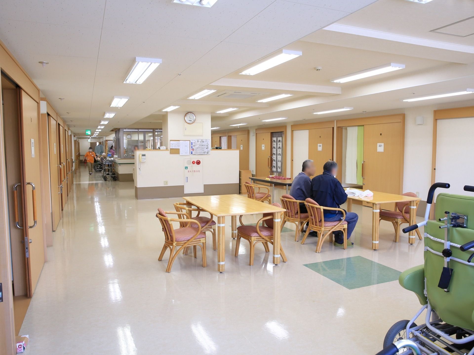 居室に囲まれた共有スペースには、木製の椅子やテーブルが複数置かれており、壁には消火設備が設置されている。