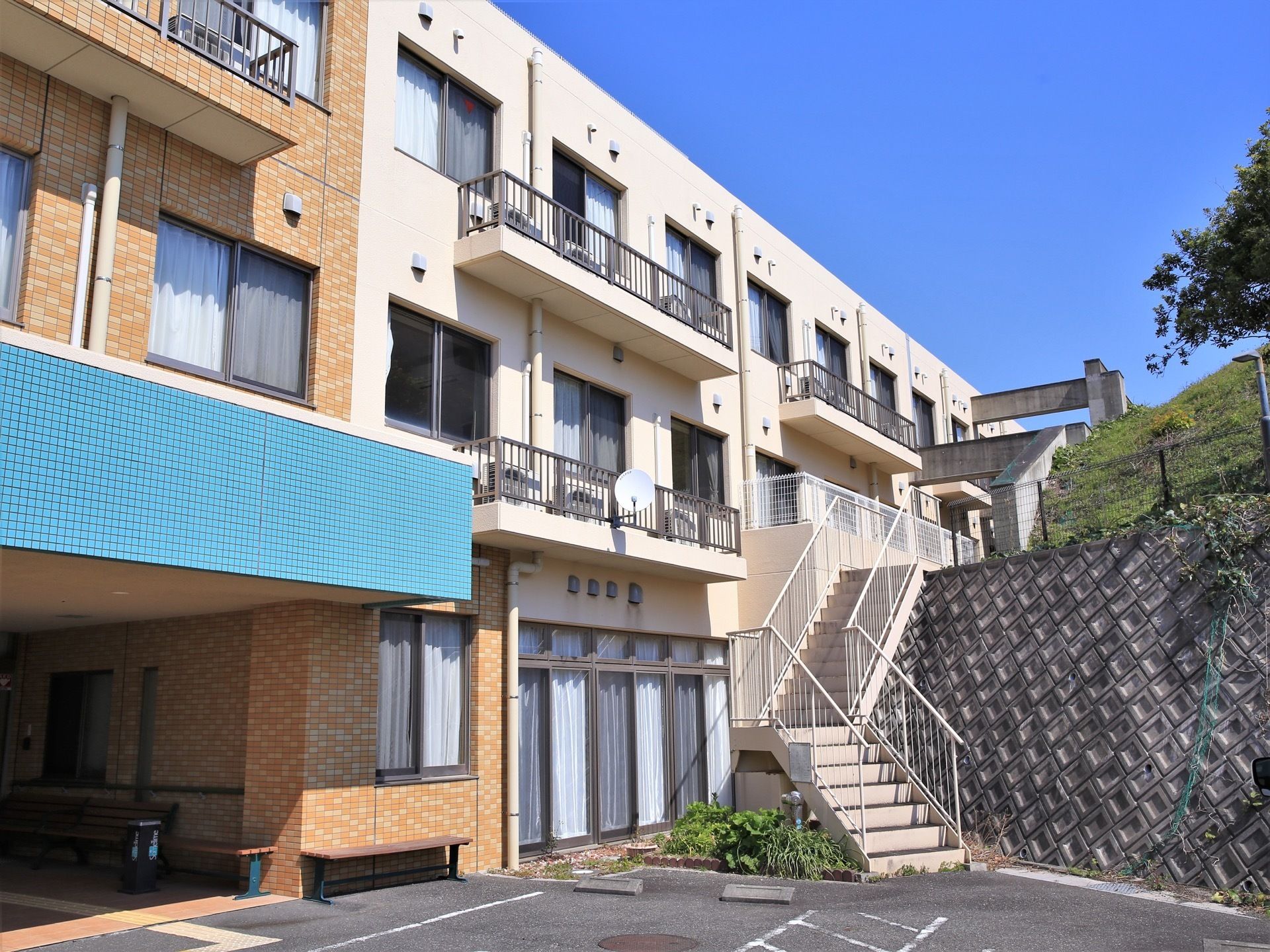 施設の外壁は、水色やベージュ色で塗装されている。居室部分にはカーテンの付いた窓と空調の室外機が設置されている。