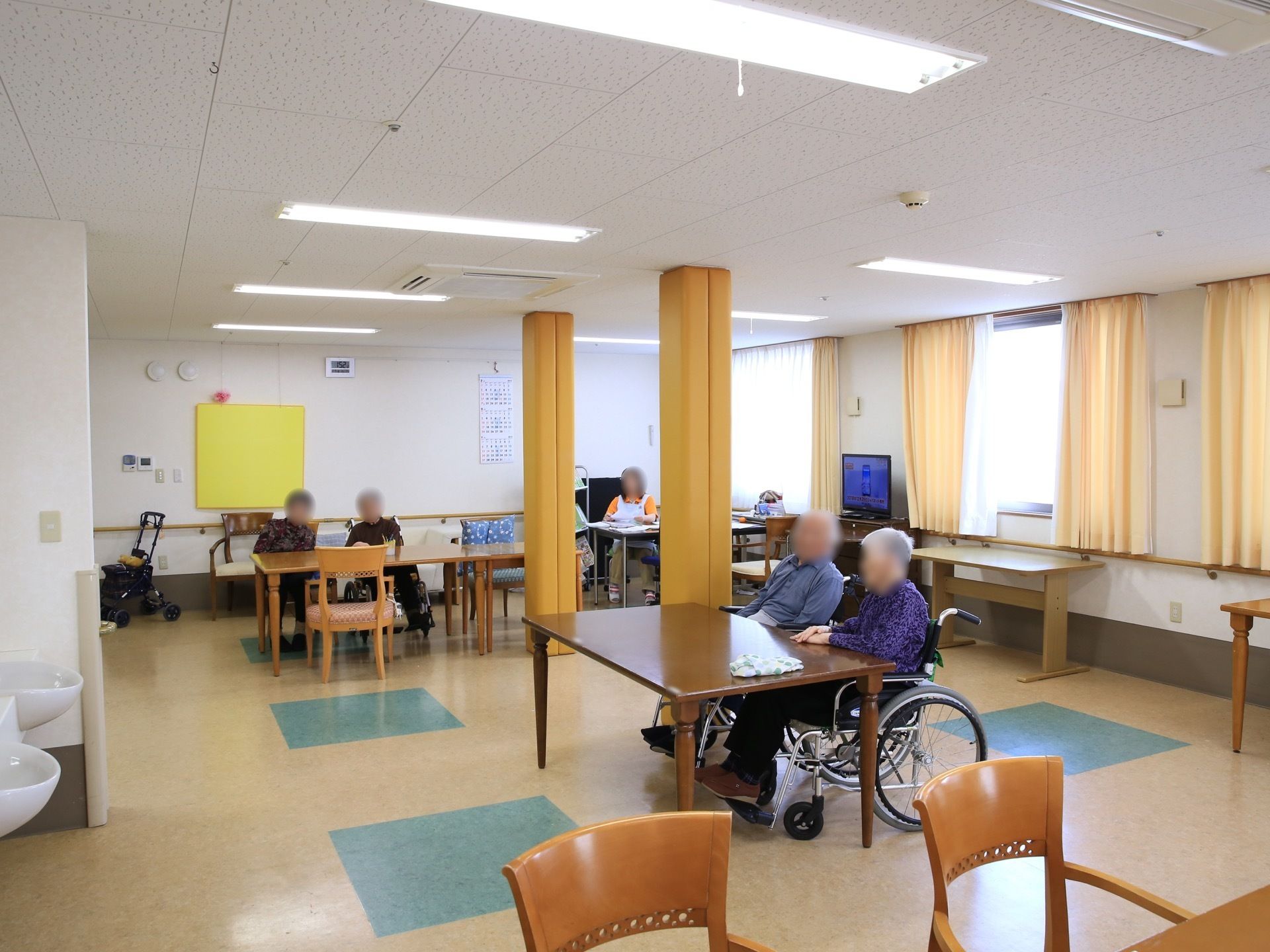 部屋には、テーブルが複数置かれており、車椅子をご利用の入居者様が並んで座っている様子である。奥には、スタッフもいる。