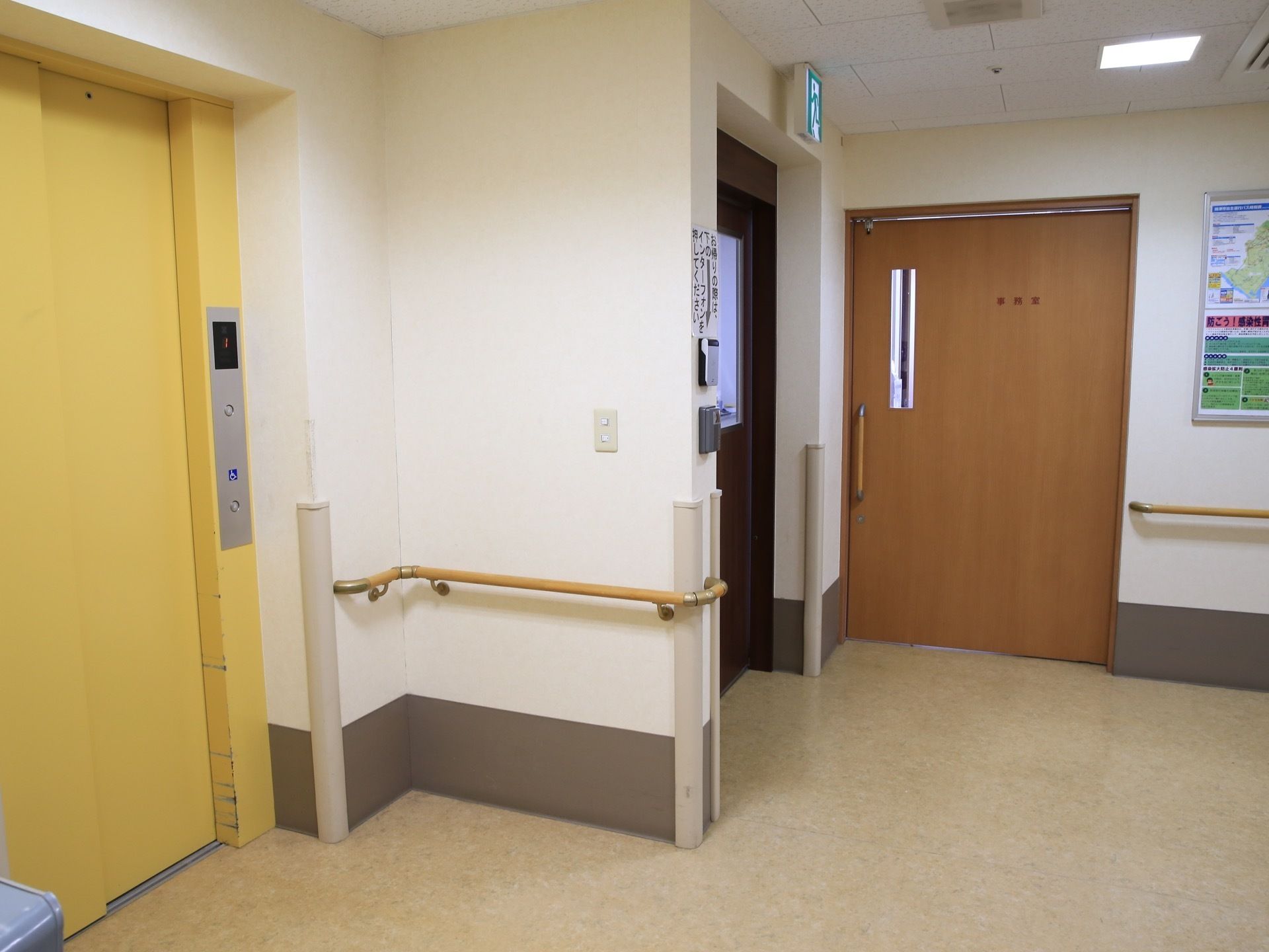 白い壁の空間の中に、黄色いエレベーターがある。エレベーターの横には非常口が設置されており、天井付近には非常口案内板が付けられている。