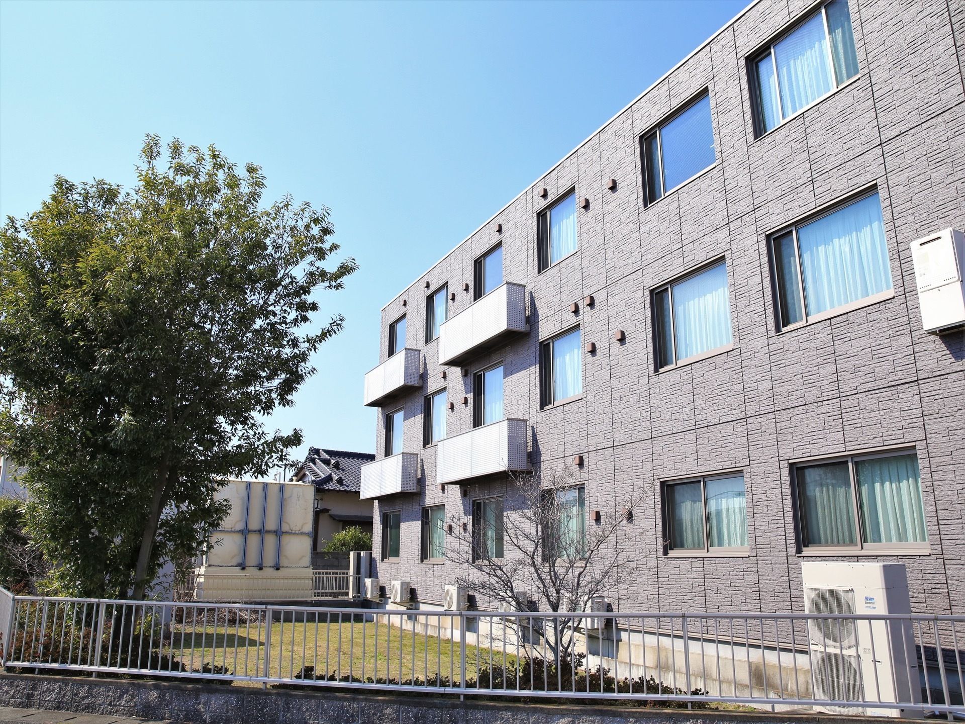 ２、３階の一部にベランダがついている、グレーの壁の３階建ての建物である。エアコンの室外機が複数置かれている。