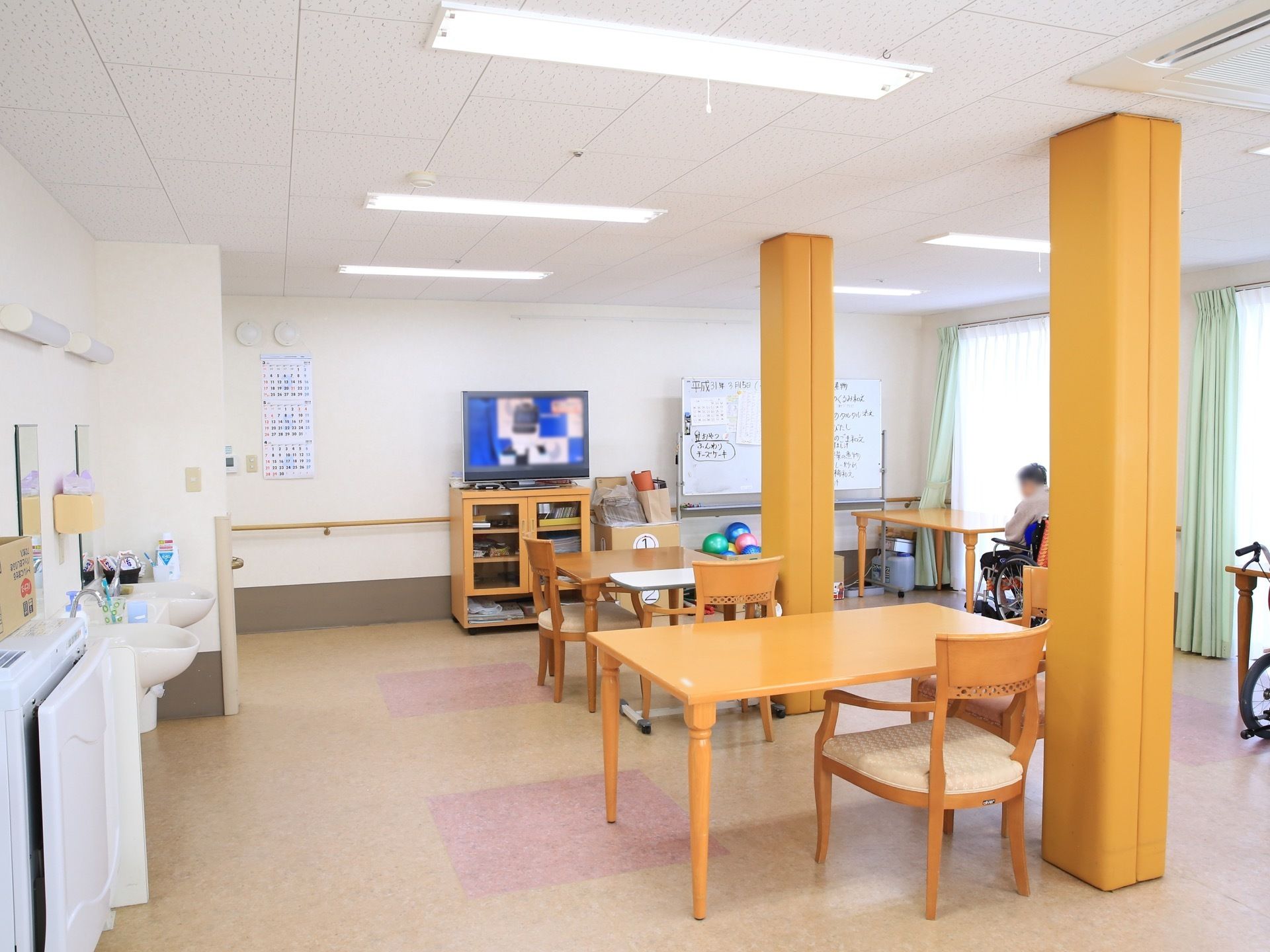白い壁、天井に、ベージュ、ピンクの床のデザインの共有スペースである。テーブルや椅子、テレビや洗面台が置かれている。