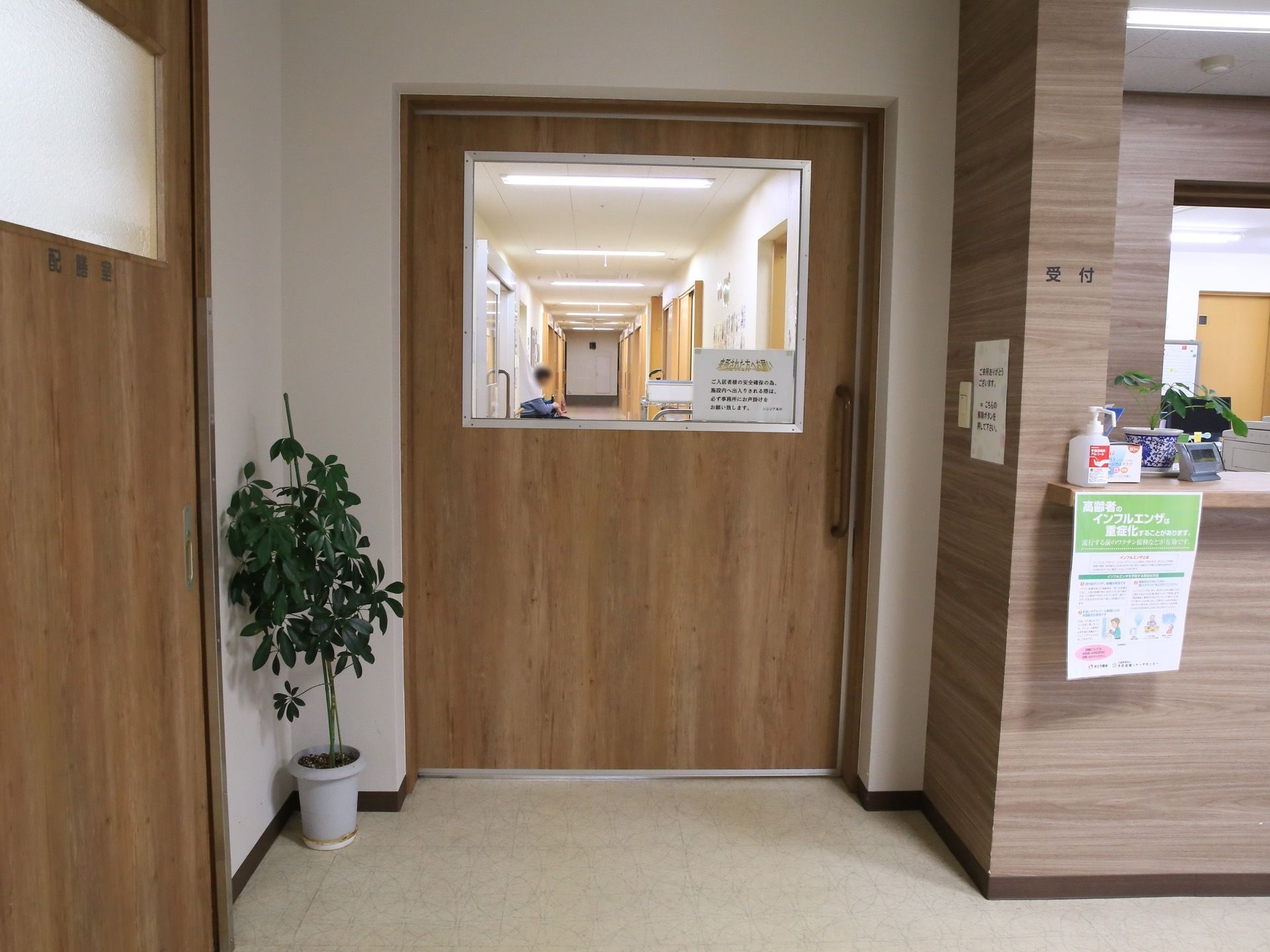 受付のカウンター横には、大きなガラス窓と長い取っ手が付いた扉がある。扉の前には観葉植物を飾っている。
