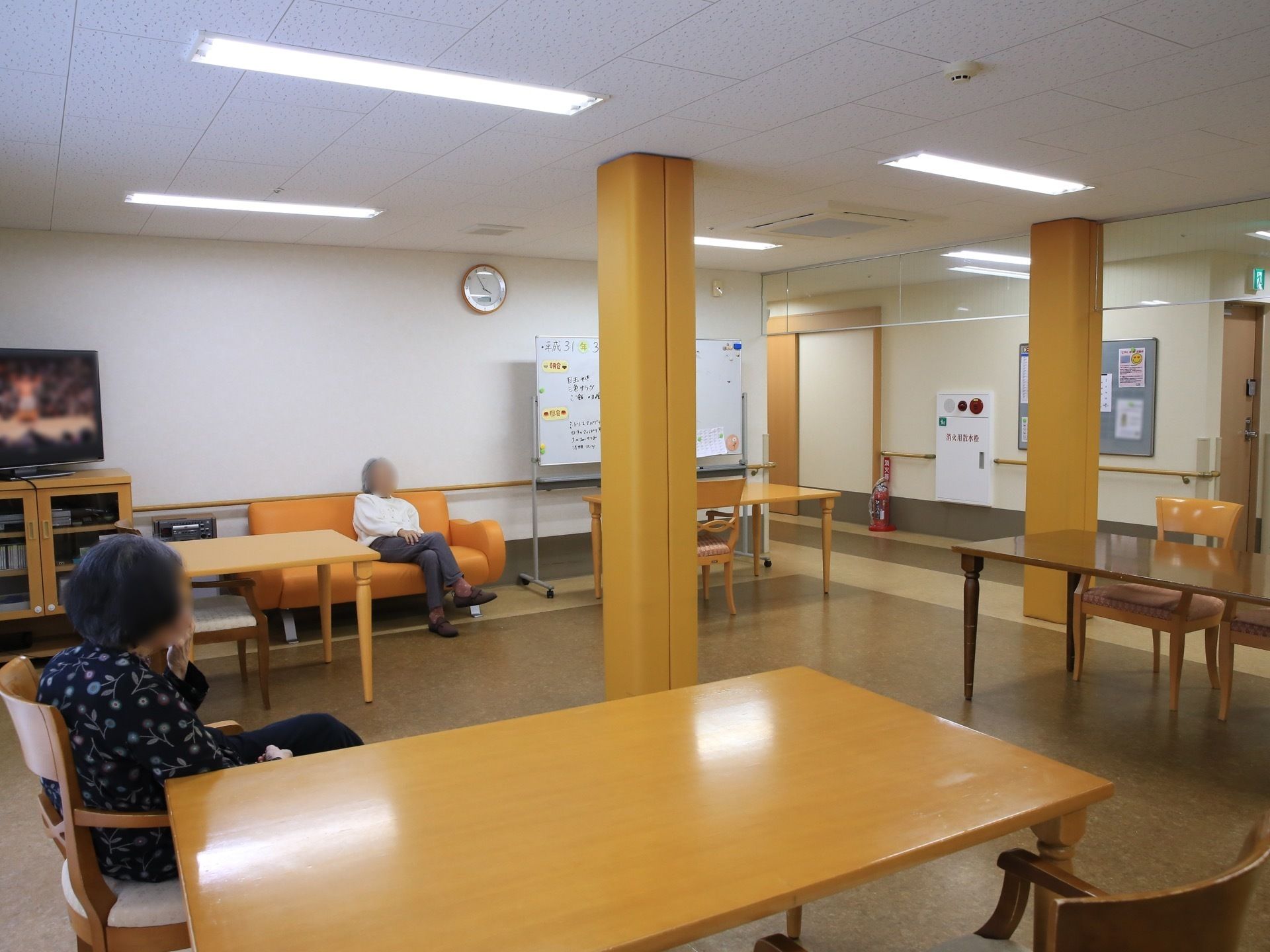 食堂の壁際にテレビが置かれている。その横には、オレンジ色のソファチェアがあり、入居者様が１名座っている。