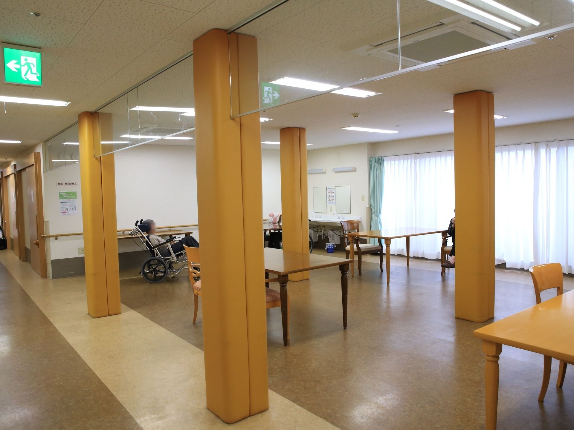 食堂には大きな窓がある。車椅子をご利用の入居者様が食堂に来ている。角が丸みを帯びた柱が４本あり広い空間である。