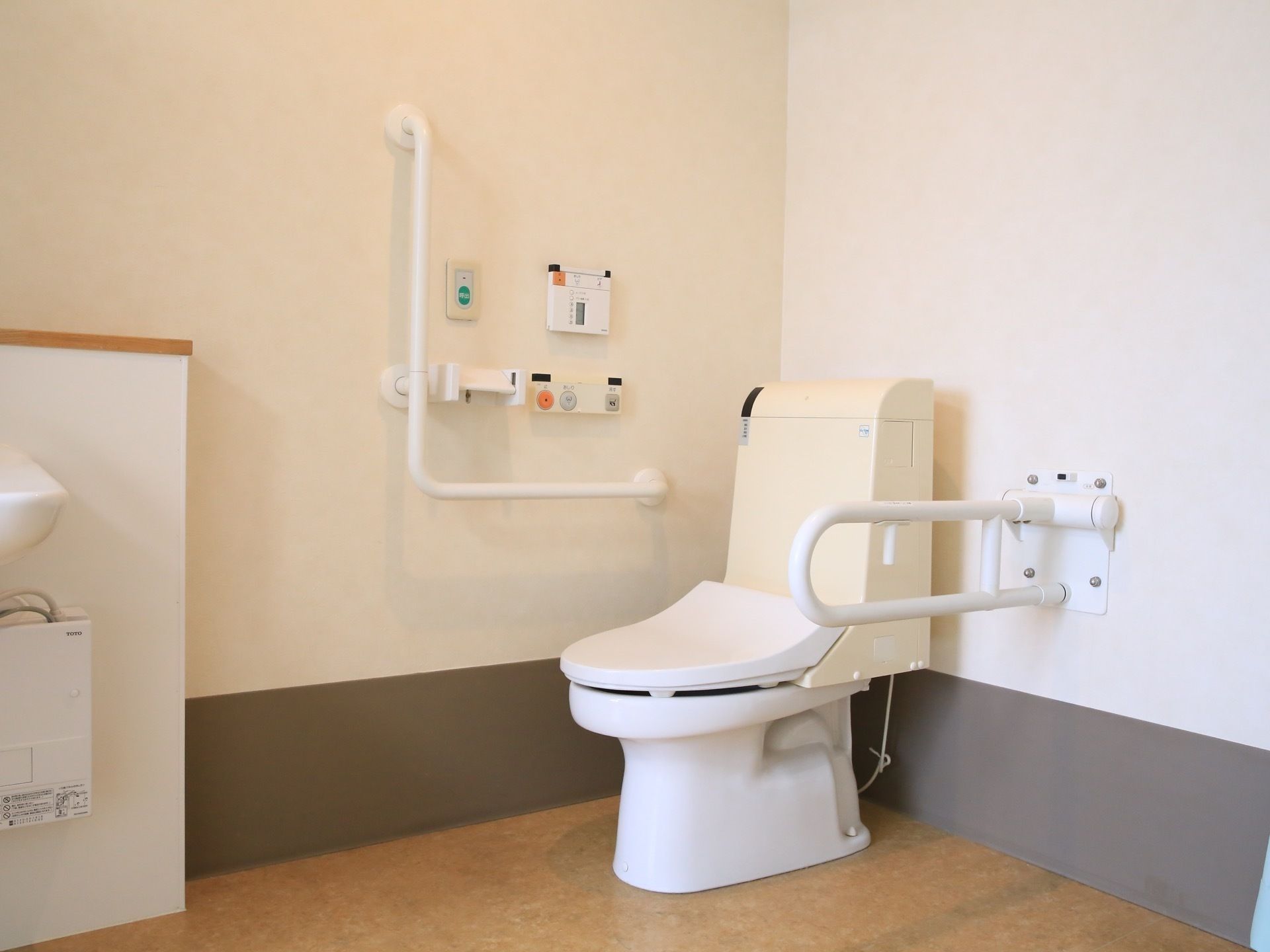 広いスペースにトイレが設置されている。可動式の手すりが便座の横にある。便座機能の操作パネルの上にナースコールボタンがついている。