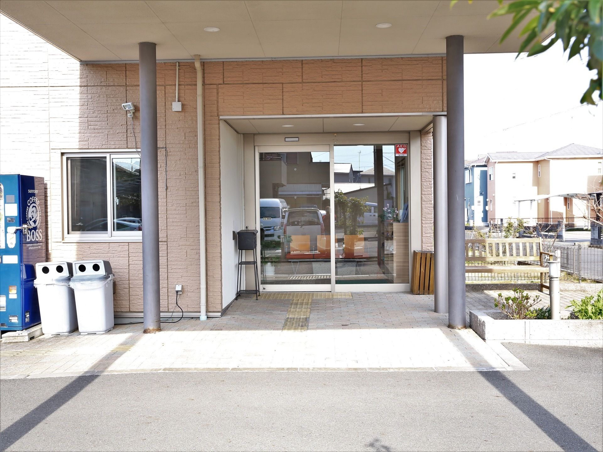 玄関前には大きなひさしがある。センサー式の照明器具がガラス窓の上に設置されている。飲料の自動販売機とくず入れが置かれている。