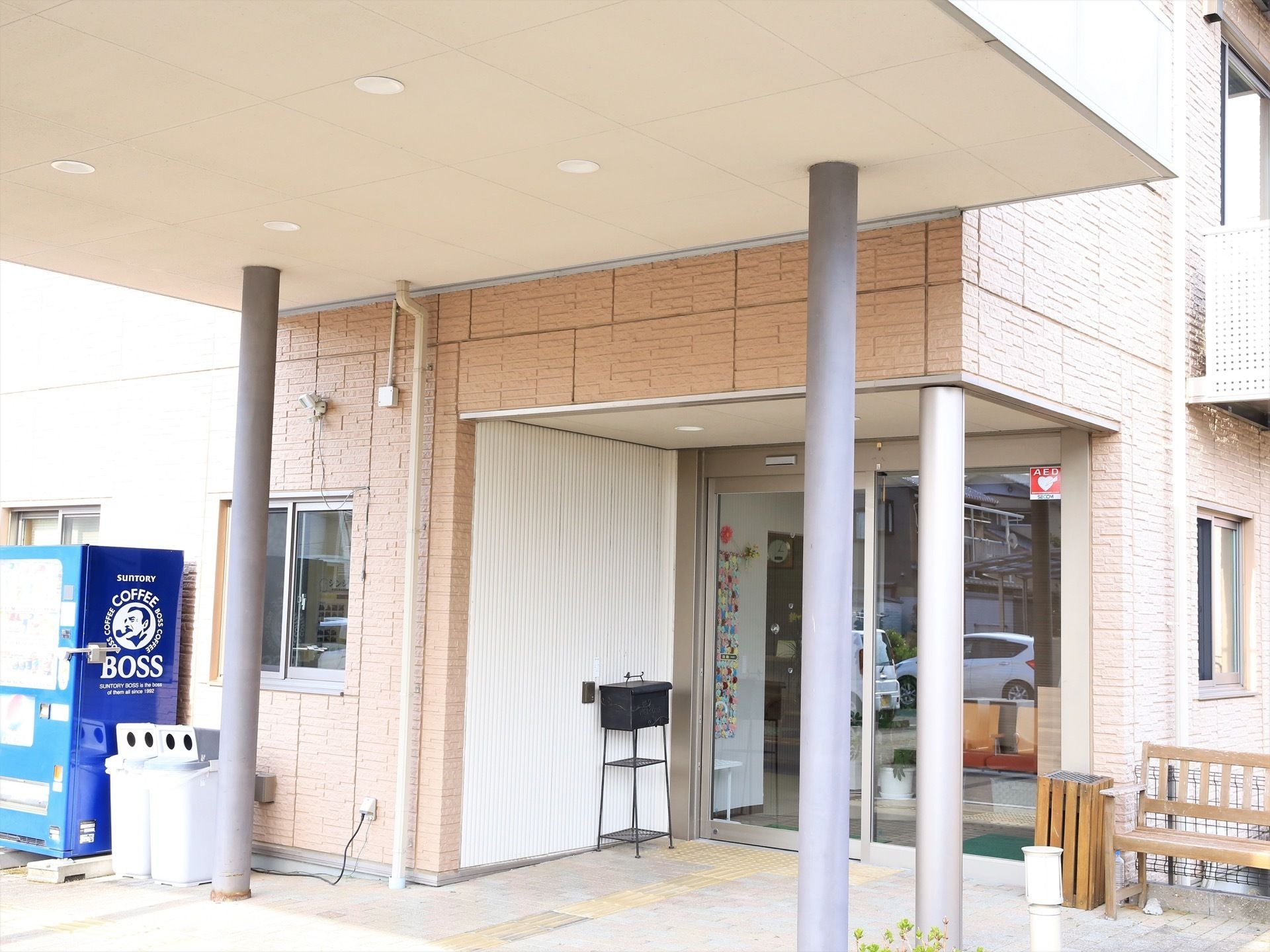 玄関の自動扉の横に、灰皿とベンチが置かれている。ひさしの左側には、飲料の自動販売機が設置されている。